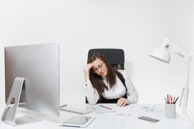 A bela mulher de negócios de cabelos castanhos cansada e cansada com dor de cabeça, sentada à mesa, trabalhando em um computador contemporâneo com monitor moderno com documentos em um escritório leve