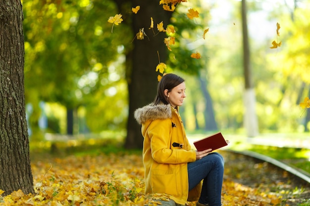 A bela mulher feliz de cabelos castanhos em um casaco amarelo e jeans senta-se sozinha no parque perto dos trilhos do bonde e lê um livro no dia quente de outono. Folhas de outono amarelas.