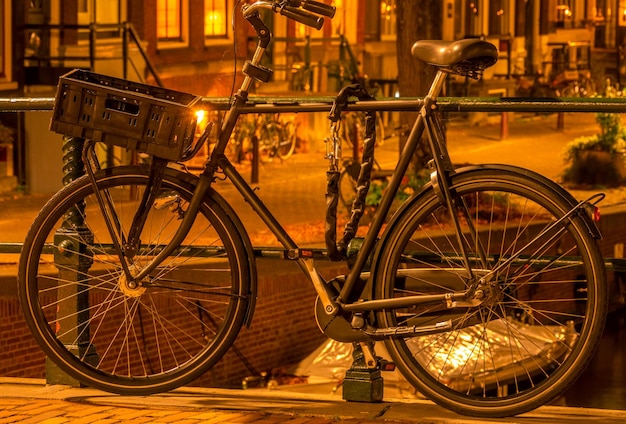 A bicicleta da holanda night amsterdam com uma cesta de bagagem está estacionada na cerca do canal e protegida com uma corrente antifurto