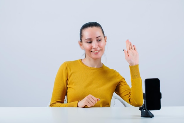 A blogueira sorridente está mostrando oi gesto sentado na frente da câmera móvel no fundo branco