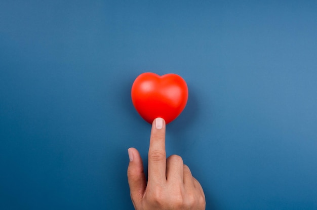 A bola de coração vermelho está apontando ou tocando com o dedo no fundo azul. Conceitos de amor, cuidado, compartilhamento, doação, bem-estar e doação, estilo minimalista.