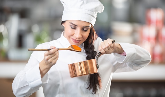 A chef profissional feminina cheira o molho com uma colher de pau, segurando uma panela na outra mão.