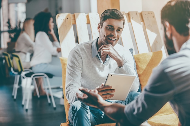 A colaboração é a chave para o sucesso. Jovem sorridente segurando um tablet digital e discutindo algo com um homem sentado à sua frente na área de descanso do escritório