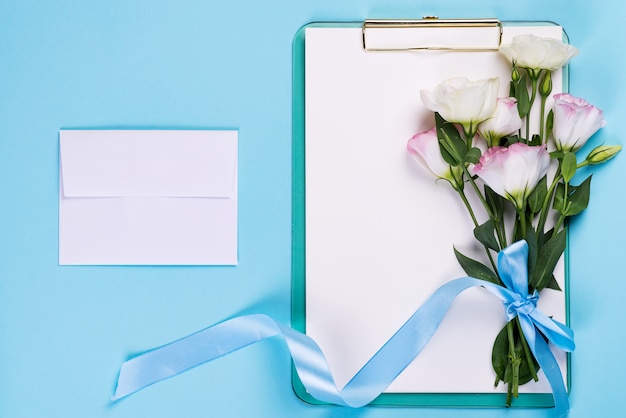 A composição mínima com um Eustoma floresce em um envelope com prancheta em um fundo azul, vista superior. Dia dos namorados, aniversário, mãe ou cartão de felicitações de casamento