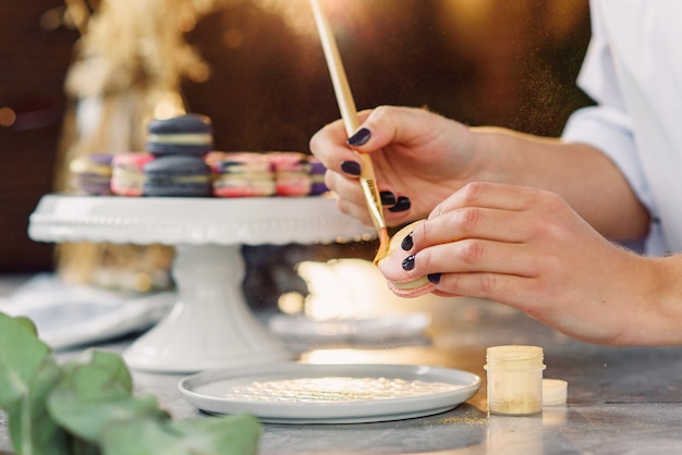 A confeiteiro profissional pinta com um pincel um farb de comida de ouro em um macaron delicioso fresco.