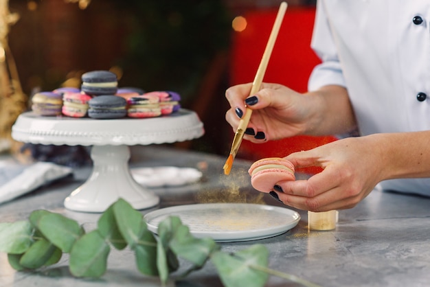 A confeiteiro profissional pinta com um pincel um farb de comida de ouro em um macaron delicioso fresco.