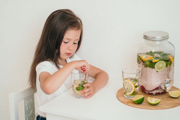 A criança bebe uma bebida com vitaminas. Limonada gelada caseira com limão, lima e hortelã. A garota se senta à mesa e bebe limonada.