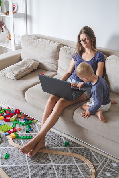 A criança brinca no sofá enquanto a mãe trabalha com o laptop, distrai e interfere no trabalho da mãe