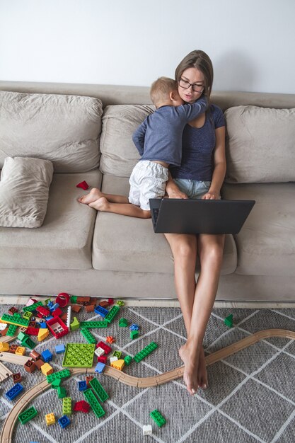 A criança brinca no sofá enquanto a mãe trabalha com o laptop, distrai e interfere no trabalho da mãe