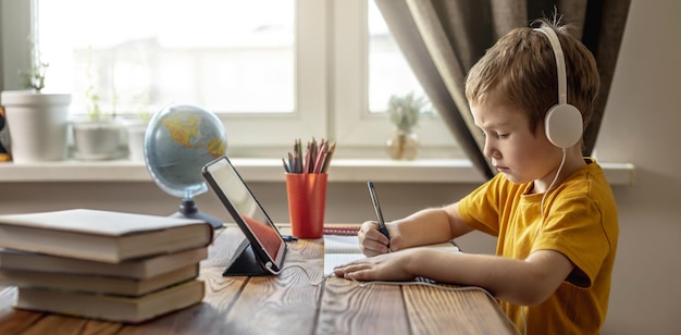 A criança é um menino em uma camiseta amarela com fones de ouvido fazendo lição de casa em um tablet Conceito de educação escolar remota