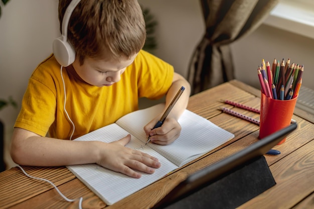 A criança é um menino em uma camiseta amarela com fones de ouvido fazendo lição de casa em um tablet Conceito de educação escolar remota