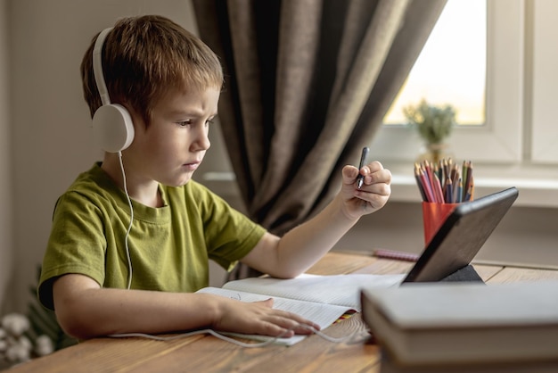 A criança é um menino em uma camiseta verde com fones de ouvido fazendo lição de casa em um tablet Conceito de educação escolar remota