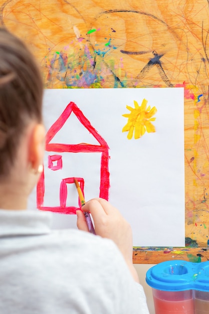 A criança está desenhando uma casa com aquarelas no cavalete.