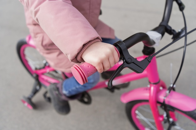 A criança está segurando o volante de uma bicicleta. Uma garota em uma jaqueta rosa está andando de bicicleta.