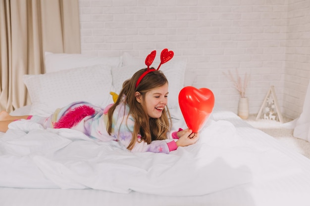 A criança está segurando um balão em forma de coração. uma garotinha deita na cama e segura um coração. Dia dos namorados