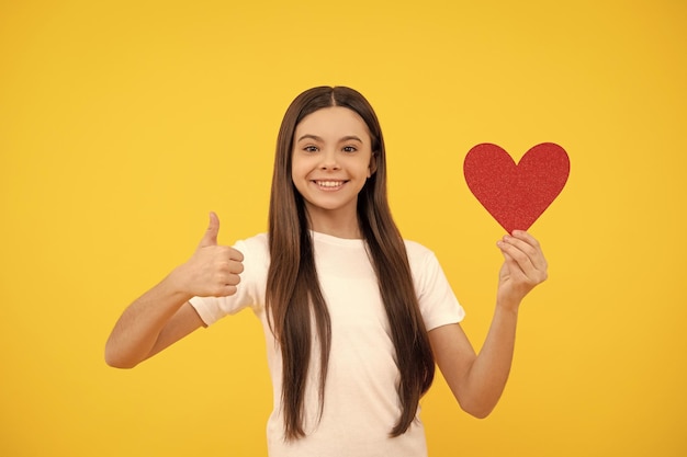 A criança feliz segura o coração do amor mostra o polegar para cima no amor de fundo amarelo