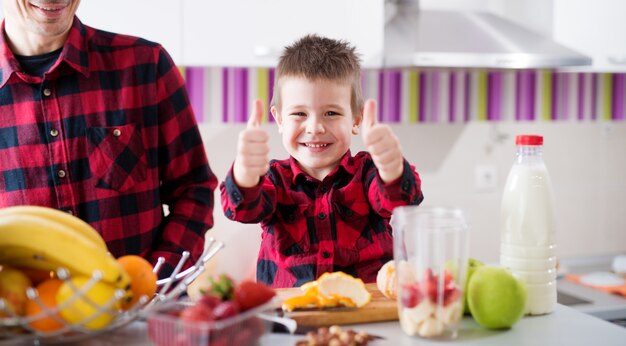 A criança orgulhosa feliz nova está aparecendo polegares ao preparar o lanche saudável da fruta com seu pai que senta-se no balcão da cozinha brilhante.