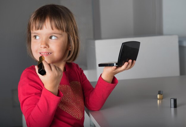 A criança pequena a menina com batom e um espelho nas mãos