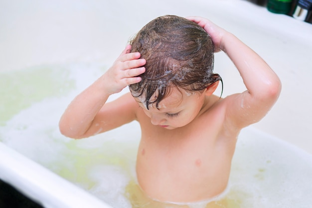 A criança pequena toma banho no banheiro e se lava a si mesmo a cabeça com o xampu