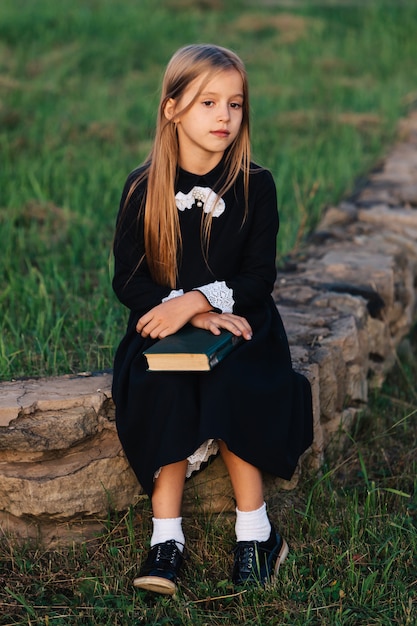 A criança se senta em um banco de pedra com um livro nas mãos e olha para longe.