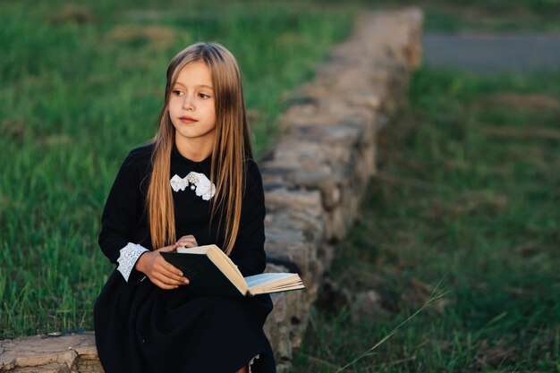 A criança se senta em um banco de pedra, segura um livro nas mãos e olha para longe.