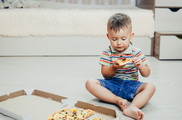 A criança senta no chão e come pizza bem apetitosa e gulosa, de bermuda e camiseta