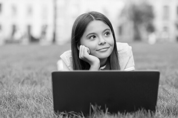 A criança sonhadora relaxa no notebook na grama verde no parque e estuda o relaxamento da educação escolar on-line