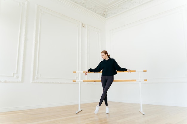 A dançarina de balé clássico tem alongamentos perto das barras de balé usa moletom preto e leggings faz exercícios em estúdio focados para baixo Instrutora certificada de coreografia