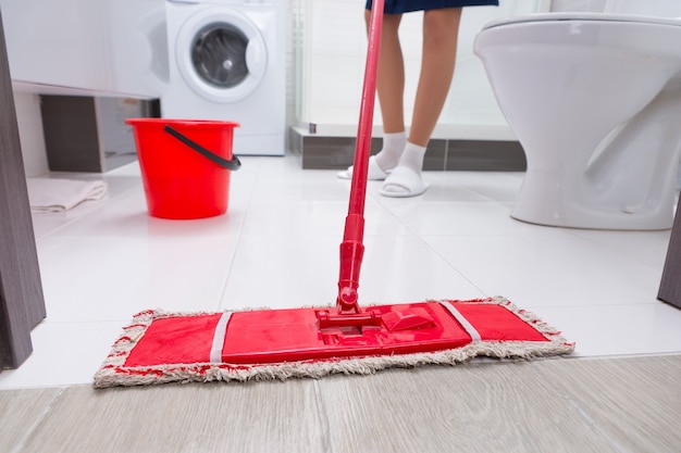 A dona de casa esfregando o chão em um banheiro com uma visão de perto de um esfregão vermelho colorido e seus pés atrás