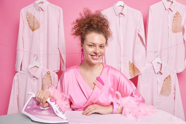 A dona de casa positiva com cabelo encaracolado vestida com manto doméstico usa ferro elétrico ocupada acariciando todas as poses de roupas de família lavadas na lavanderia se sente feliz por ter um dia agitado. Conceito de rotinas de limpeza