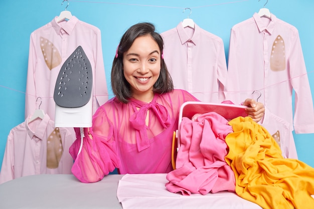 A dona de casa sorridente e autoconfiante segura o ferro elétrico a vapor fica satisfeita quando está passando roupas com roupas passadas. A dona de casa feliz tem muito trabalho a fazer