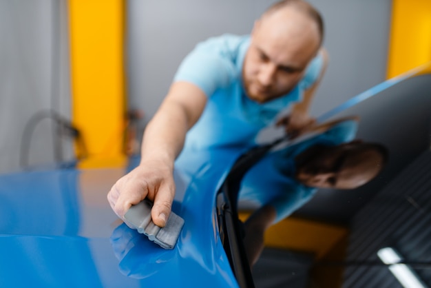 A embalagem de carro masculino com rodo instala uma película protetora de vinil ou filme no capô. Trabalhador faz detalhamento de automóveis. Revestimento de proteção de pintura automotiva, ajuste profissional