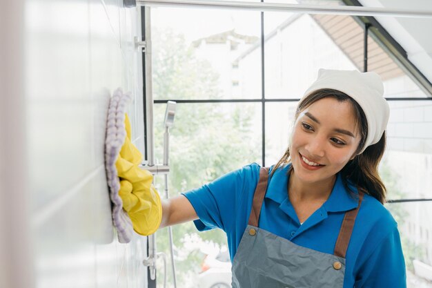 A empregada doméstica, usando luvas de proteção, limpa a poeira dos azulejos de cerâmica com um pano de microfibras no banheiro. Sua limpeza de rotina enfatiza a pureza e a higiene na limpeza comercial.