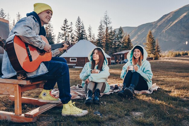 A empresa está descansando na madeira do acampamento florestal. cara sentado na cadeira tem um violão nas mãos e toca uma melodia. as meninas estão sentadas ao lado dele e bebendo chá apreciando a atmosfera.