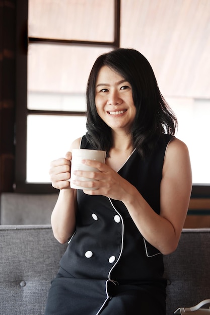 A empresária asiática sorridente está bebendo e relaxando com uma xícara de café no espaço de trabalho no escritório