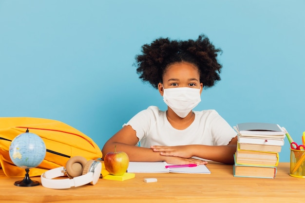 A estudante afro-americana em uma máscara senta-se em uma mesa na sala de aula em um fundo azul De volta à escola e ao conceito covid19