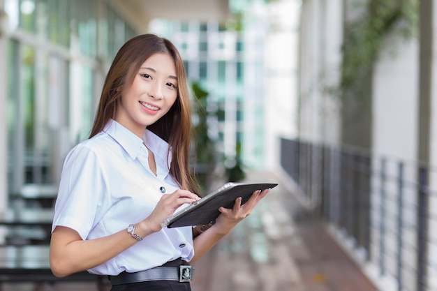 A estudante tailandesa asiática de uniforme está sorrindo alegremente e com confiança enquanto usa o tablet