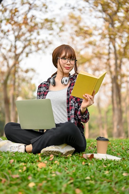 A estudante universitária asiática focada senta-se na grama, lê um livro e usa seu laptop