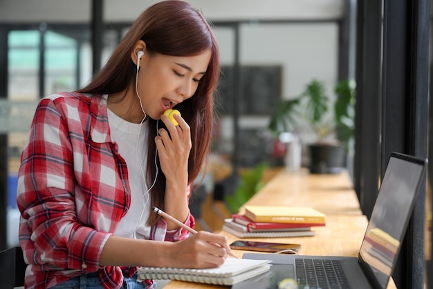 A estudante universitária asiática senta-se no café fazendo sua casa ouvindo música e comendo macaron