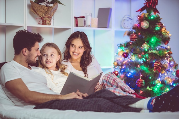 A família deitou na cama e leu um livro perto da árvore de natal. hora da noite