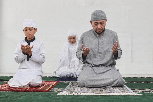 A família muçulmana asiática está sentada e rezando dua juntos na mesquita