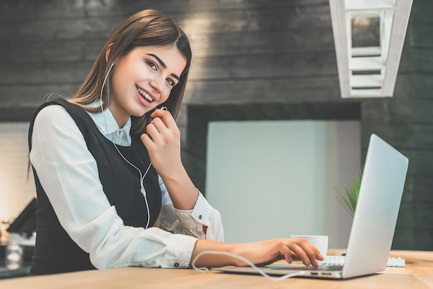 A feliz empresária em fones de ouvido sentada com o laptop moderno à mesa