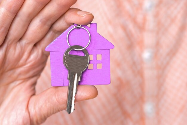 A figura de uma mini casa roxa com as chaves está nas mãos de um homem em um fundo bege