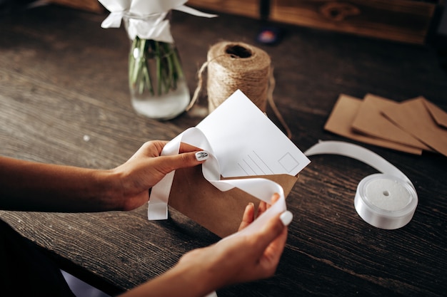 A florista está embalando um cartão postal em um envelope com fita branca sobre a mesa de madeira.