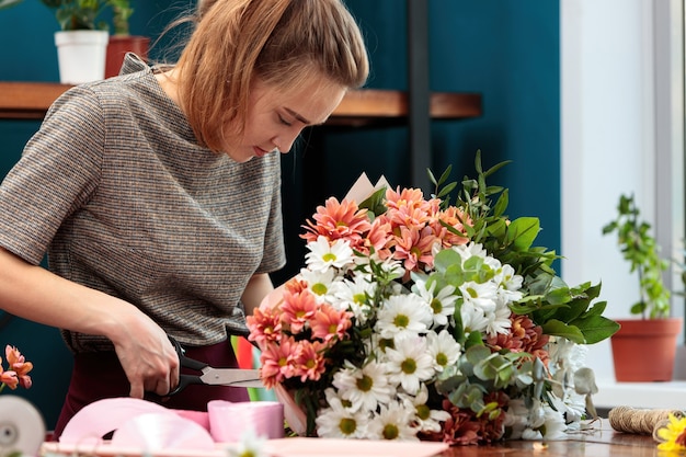 A florista faz um buquê de crisântemos. uma jovem adulta corta uma fita para decoração com uma tesoura.