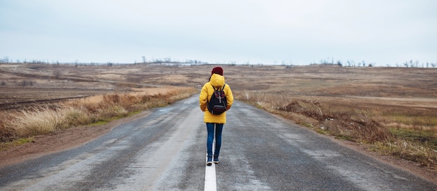 A foto de grande angular de uma turista feminina com uma mochila vestindo jaqueta amarela e chapéu vermelho caminha na estrada.