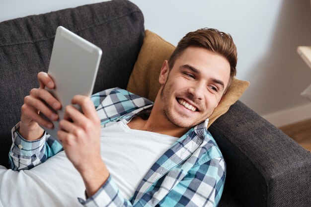 A foto de um homem sorridente com cerdas, vestido com uma camisa e uma gaiola estampada, encontra-se no sofá em casa e usando o computador tablet.