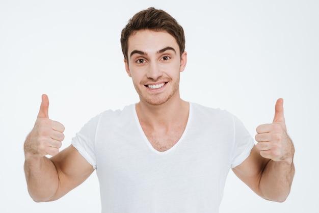 A foto de um jovem feliz vestido com uma camiseta branca em pé sobre um fundo branco faz o gesto de polegares para cima.