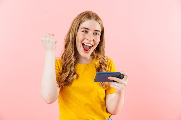 A foto de uma jovem garota emocional gritando isolada sobre a parede rosa usando o telefone celular faz o gesto do vencedor.