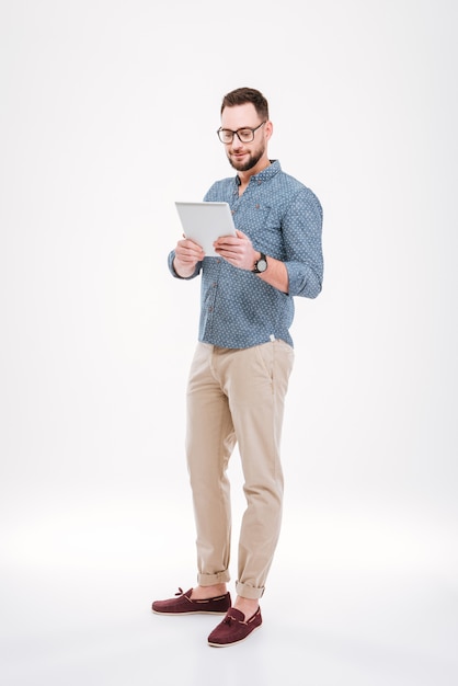 A foto de vidros farpados atrativos do homem barbudo vestiu-se na camisa azul usando o tablet pc isolado sobre a parede branca.